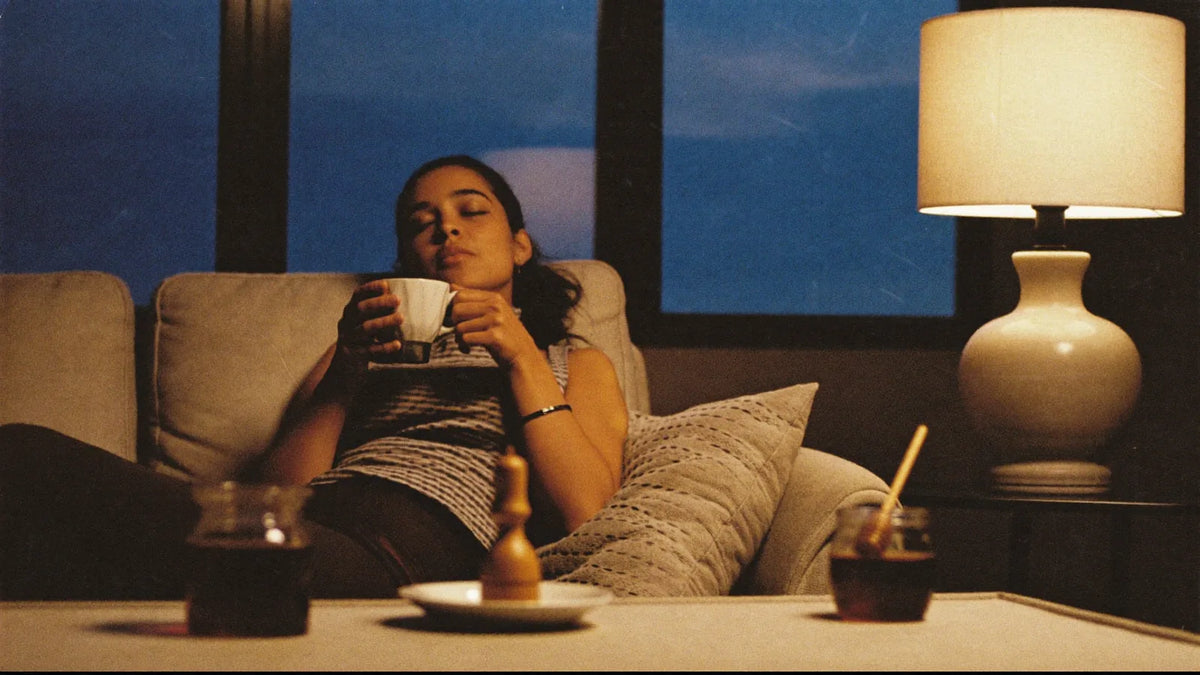 A woman sunk into the sofa at dusk with a warm mug of tea, honey jar on the table — calm and content