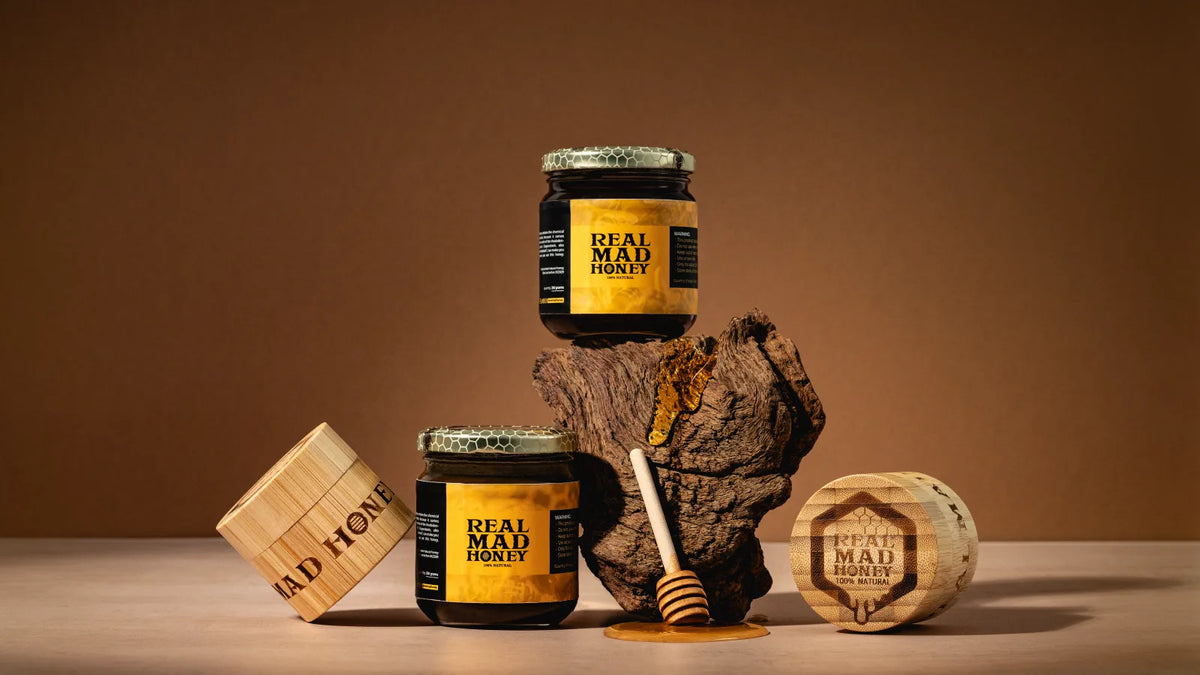 A jar of Real Mad Honey with its engraved bamboo lid, a wooden dipper and honeycomb, on a warm studio backdrop